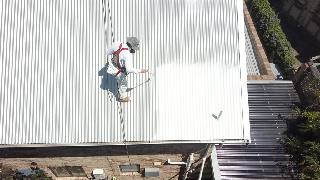 A worker conducting commercial roof repairs in Perth, painting a white metal roof.
